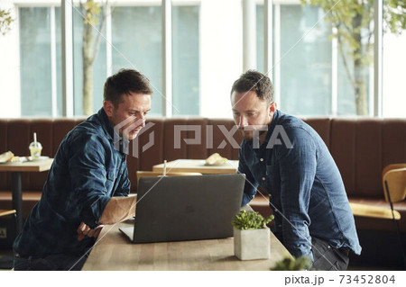 Two men seated in a cafe looking at a laptop screen 73452804