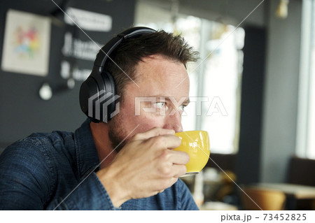 Man seated in a cafe wearing headphones, drinking coffee 73452825