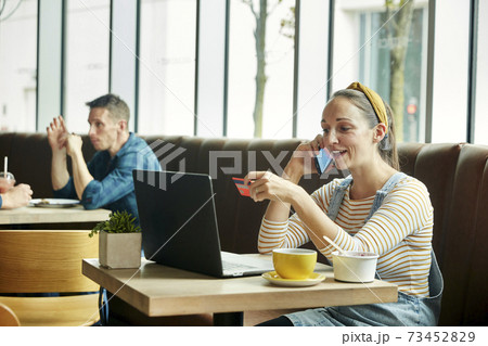 Woman seated in a cafe using a laptop and talking on a smart phone 73452829