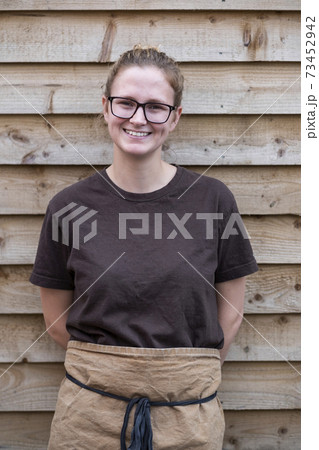 Portrait of female barista with blond hair and glasses, wearing brown apron, leaning against wooden wall, smiling at camera. 73452942