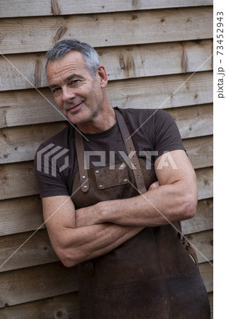Portrait of male barista with short grey hair, wearing brown apron, arms folded, leaning against wooden wall. 73452943
