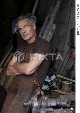 Portrait of male barista with short grey hair, wearing brown apron, arms folded, looking at camera. 73452946