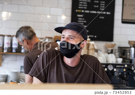 Male barista wearing black baseball cap and face mask working behind counter. 73452953