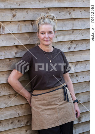 Portrait of waitress wearing brown apron, leaning against wall, smiling. 73452963