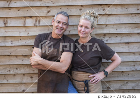 Waiter with short grey hair and waitress with blond hair, both wearing brown aprons, leaning against wooden wall, smiling at camera. 73452965