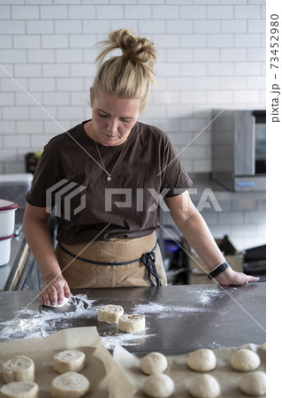 Blond woman wearing brown apron standing in a kitchen, baking danish pastries. 73452980