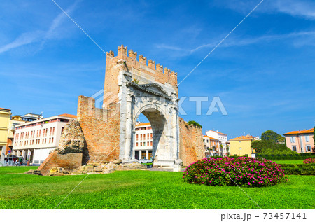 Ruins of ancient brick wall and stone gate Arch of Augustus Arco di Augusto, green lawn with bush of flowers in old historical touristic city centre Rimini 73457141