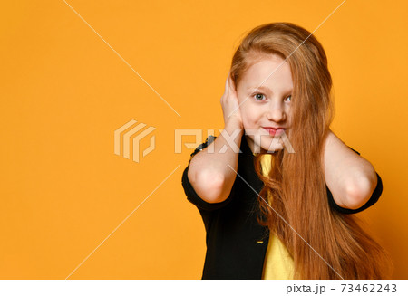Red-headed teenage kid in black jacket and yellow t-shirt. She closed her ears with hands, posing on orange background. Close up Red-headed teenage kid in black jacket and yellow t-shirt. She closed her ears with hands, posing on orange background. Close up 73462243