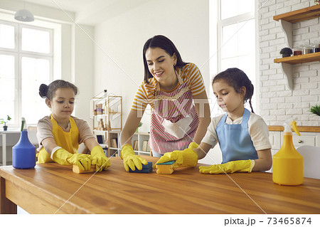 Happy little daughters helping their mom wash the table with sponges and detergent Happy little daughters helping their mom wash the table with sponges and detergent 73465874