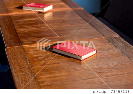 Untitled red hardcover book on a wooden brown table lit by the sunlight of a empty street cafe, nobody closeup. Untitled red hardcover book on a wooden brown table lit by the sunlight of a empty street cafe, nobody closeup. 73467113