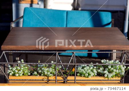 Designer flowerpot made of iron tubular frame with green flowers and white blossoms at the brown wooden table with blue chair of an outdoor cafe, close up nobody. 73467114