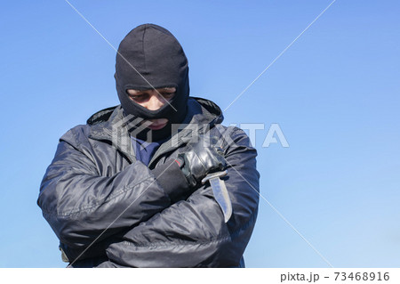 a sad, thoughtful bandit stands in a black mask and with a knife against the blue sky 73468916