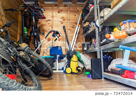 Suburban home wooden storage utility unit shed with miscellaneous stuff on shelves, bikes, exercise machine, ladder, garden tools and equipment. Messy and chaos at house yard barn. Organization order 73472014