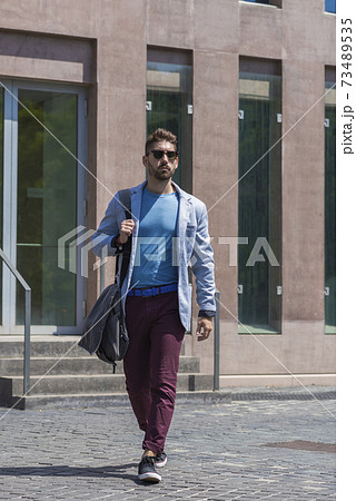 Young businessman walking next to office buildings while holding a shoulder bag outdoors Young businessman walking next to office buildings while holding a shoulder bag outdoors 73489535