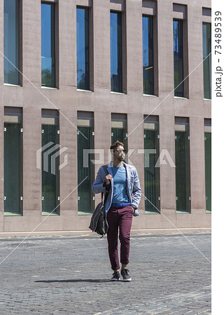 Young businessman walking next to office buildings while holding a shoulder bag outdoors 73489539