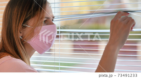 Young woman in pink home made cotton virus face mask, looking through window blinds outside. Quarantine or stay at home during coronavirus covid-19 outbreak Young woman in pink home made cotton virus face mask, looking through window blinds outside. Quarantine or stay at home during coronavirus covid-19 outbreak 73491233