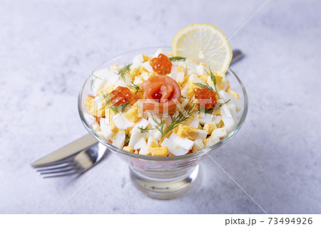 Layered salad with salted trout (salmon), caviar, eggs and vegetables. Traditional dish, one portion in a glass goblet on a gray background. Close-up. 73494926