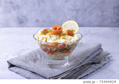 Layered salad with salted trout (salmon), caviar, eggs and vegetables. Traditional dish, one portion in a glass goblet on a gray background. Close-up. 73494929