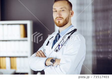 Red-bearded doctor standing straight in clinic near his working place. Portrait of physician. Medicine and healthcare concept Red-bearded doctor standing straight in clinic near his working place. Portrait of physician. Medicine and healthcare concept 73517403