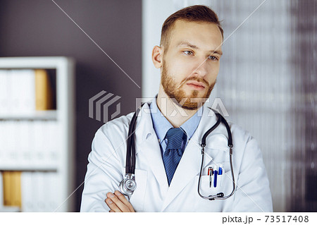 Red-bearded doctor standing straight in clinic near his working place. Portrait of physician. Medicine and healthcare concept 73517408