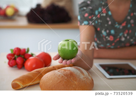 Close-up of female hand holding green apple in kitchen interiors. Many vegetables and other meal at glass table are ready for been cooked soon 73520539