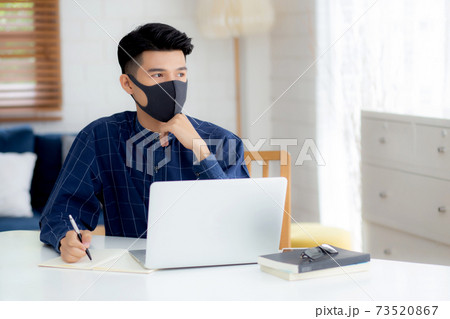 Young business man in face mask planning and working from home on laptop computer on desk. 73520867