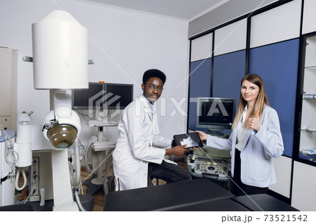 Portrait of smiling African man doctor, holding digital tablet with ultrasound scan, posing to camera, while consulting his female patient in modern clinic. Woman patient shows thumb up 73521542