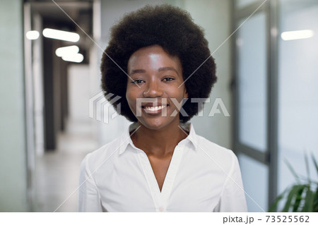 Close up face portrait of pretty smiling black young woman with afro hair, wearing white shirt, looking at camera, while standing inside modern office building. Businesswoman indoor portrait Close up face portrait of pretty smiling black young woman with afro hair, wearing white shirt, looking at camera, while standing inside modern office building. Businesswoman indoor portrait 73525562