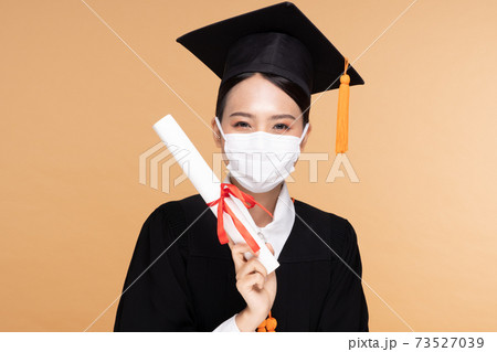 Attractive Asian woman graduated in cap and gown wear medical mask smile so proud and happiness due Virus outbrake and COVID-19 virus flu pandemic on Beige background,New normal of graduation concept 73527039