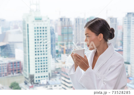 Asian woman in pampering spa resort luxury hotel drinking tea relaxing wearing bathrobe looking at view from bedroom window. Asian travel lady enjoying room service 73528285