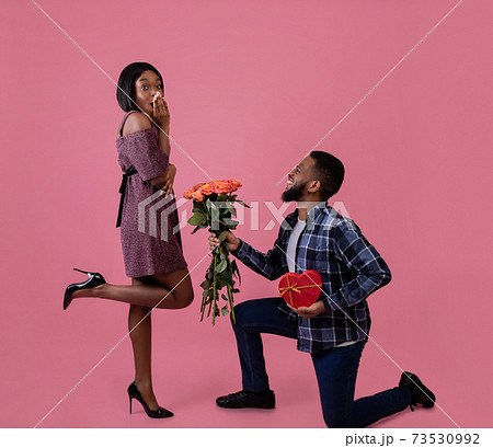 Passionate black man giving his sweetheart bouquet of roses and heart shaped gift for Valentine's Day on pink background 73530992