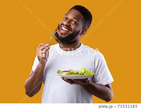 Cheerful Black Guy Eating Salad Enjoying Healthy Meal, Yellow Background Cheerful Black Guy Eating Salad Enjoying Healthy Meal, Yellow Background 73531165