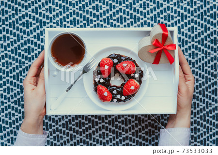 Top view female hands holding romantic meal for Lover. A cup of tea, bagel with jam decorated with strawberries and heart-shaped gift box on the white wooden tray. Simple surprise for Valentine's day 73533033