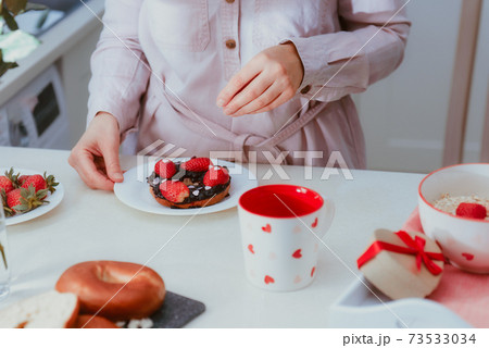 Woman decorating sweet bagel with jam with strawberries and heart-shaped candies. Preparing surprise breakfast for lover on Valentines' day. Simple festive food ideas. Copy space 73533034