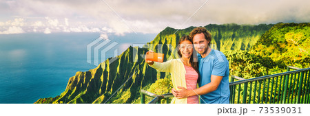 Travel selfie couple at Napali Kauai Hawaii banner. Young tourists at Na Pali Coast lookout taking mobile phone selfie smartphone photo. Travel people at hawaiian landscape, panorama crop 73539031