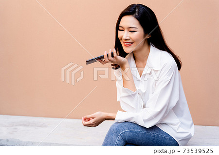 Young smiling Asian girl having conversation on smartphone while sitting on concrete bench with a desert sand colored wall in background wearing white shirt and jeans 73539525