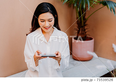 Asian woman playing games on smartphone while sitting on a concrete bench surrounded with pots of green plants with a desert sand colored wall in her background 73539526