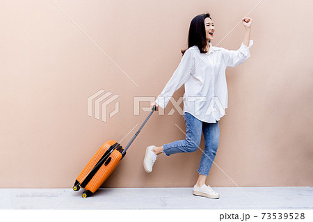 Young Asian girl happy while towing her bright orange suitcase with right hand and left hand fist bumping in the air on concrete floor and desert sand colored wall in her background 73539528