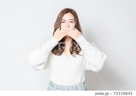 白い壁の前に立って、手で口を隠す若い女性 73539645