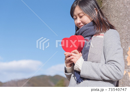青空の下でハートの箱を持って木に寄りかかり思いを巡らす女性 73540747