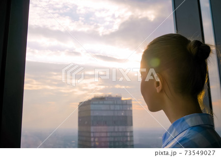 Woman looking at cityscape through window of skyscraper - sun lens flare Woman looking at cityscape through window of skyscraper - sun lens flare 73549027