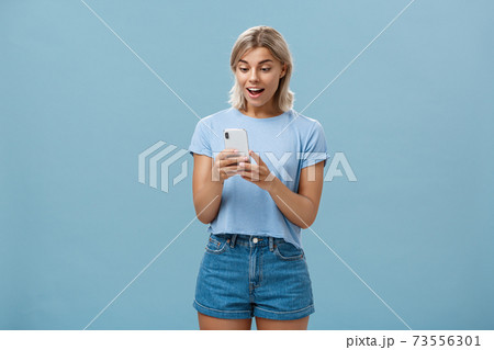 Entertained charming young european female with blond hair in casual outfit holding smartphone being satisfied with great camera and photos gasping and gazing amazed at screen over blue wall 73556301
