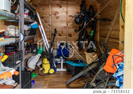Suburban home wooden storage utility unit shed with miscellaneous stuff on shelves, bikes, exercise machine, ladder, garden tools and equipment. Messy and chaos at house yard barn. Organization order 73558332