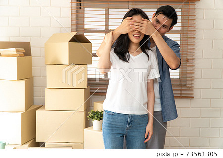 Smiling young Asian happy couple close girlfriend eyes for a surprise at moving day in their new home after buying real estate. Concept of starting a new life for a newly married couple. 73561305