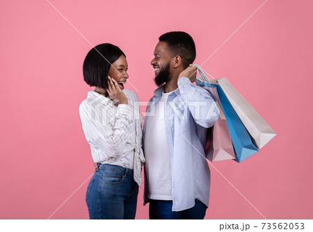 Happy young black man with his sweetheart holding shopping bags, purchasing gifts for lovers day over pink background 73562053