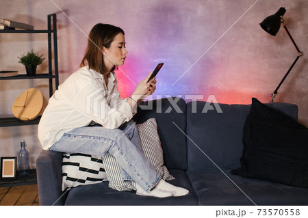 Young asian woman sitting on a sofa in a cozy room looks at the phone 73570558