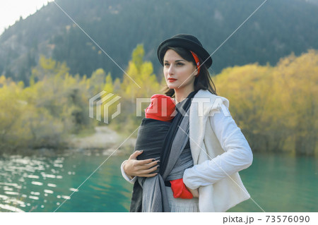 Beautiful young mother with her infant baby in sling outdoor. Mother is carrying her child and travel in autumn mountain lake. Babywearing concept 73576090