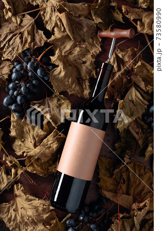 Bottle of red wine with corkscrew. On a table dried vine leaves and blue grapes. 73580990