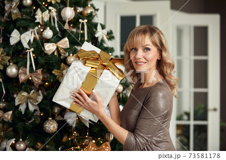 Young happy beautiful woman with gift boxes sits near Christmas tree in the room of the house. Merry Christmas and New Year. 73581178