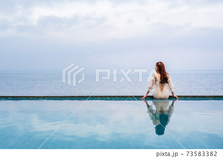 woman sitting on edge of swimming pool and looking to the sea 73583382